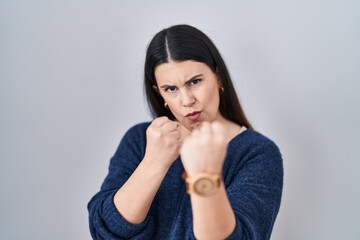 Wall Mural - Young brunette woman standing over isolated background ready to fight with fist defense gesture, angry and upset face, afraid of problem