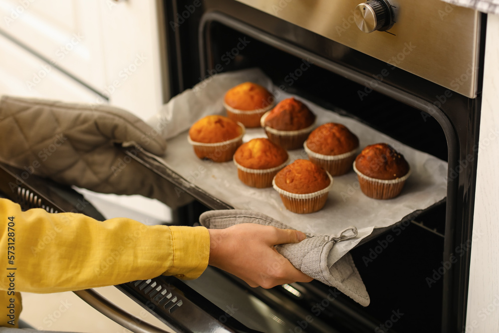 Young woman taking tray with cupcakes from oven in kitchen, closeup
