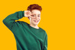 Portrait of cute friendly positive red-haired boy in green hoodie showing two fingers near his face showing v-sign. Smiling funny cheerful boy with fallen out baby teeth on isolated yellow background.