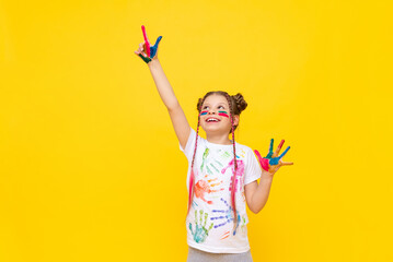 Wall Mural - A beautiful little girl with stained hands in paint points her fingers in different directions. A child in paint on a yellow background.