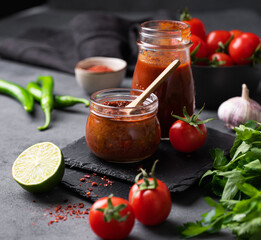 Wall Mural - Hot sauce adjika. Homemade appetizer with peppers and tomatoes in a jar on a dark background with vegetables and herbs. Rustic style.