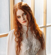 Portrait fantasy beauty red-haired woman green eyes looking at camera. White old style vintage dress. Stads by window waiting love. Curly red hair. Redhead fashion model sexy girl princess beauty face