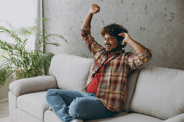 Wall Mural - Side view fun young Indian man wears casual clothes headphones listen to music sits on grey sofa couch stay at home hotel flat rest relax spend free spare time in living room indoor. Lounge concept.