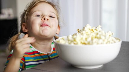 Wall Mural - Little girl is eating popcorn in home kitchen. Focus on hand taking popcorn