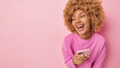 © wayhome.studio  - Horizontal shot of optimistic woman poses with phone looks aside smiles broadly dressed in casual jumper enjoys using modern technologies isolated over pink background copy space for your promo