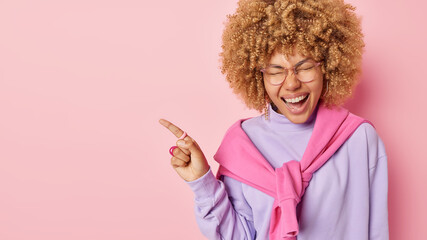 Wall Mural - Cheerful optimistic European woman with curly hair points index finger on blank space exclaims loudly demonstrates something awesome wears spectacles and jumper isolated over pink background.