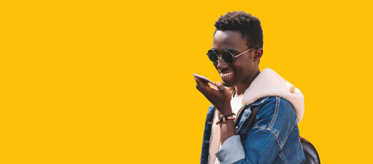 Wall Mural - Portrait of happy smiling young african man holding smartphone using voice command recorder, assistant or takes calling, looking at phone isolated on yellow background