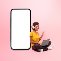 Glad young black curly female in wireless headphones with computer, wave hand near big phone with empty screen