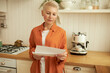 © Anatoliy Karlyuk - Horizontal portrait of concerned mature elderly lady with short haircut reading negative postal letter of notice standing against kitchen counter, standing with serious facial expression