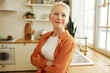 Indoor image of attractive happy confident mature female house owner standing in kitchen leaning against counter with big window, looking at camera with crossed hands and positive facial expression
