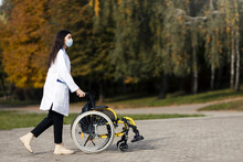 Caring Young Doctor In A Protective Mask Carries A Wheelchair. A Young Nurse Is Carrying A Cart Outdoors.