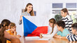 Preteens pupils looking at national flag of while learning different cultures with smiling young female teacher in schoolroom