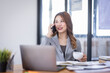 © David - Smiling young Asian woman sitting and call phone device, looking at laptop, checking modern apps, texting messages, browsing internet doing shopping relaxing at home.