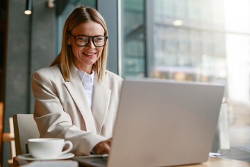 Wall Mural - Smiling businesswoman freelancer working on laptop while sitting in cafe near window