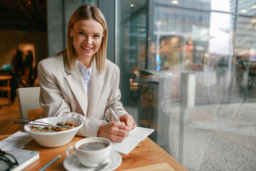Wall Mural - Attractive businesswoman having lunch and making notes while working in cafe sitting near window
