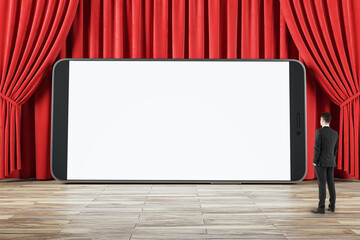 Poster - Businessman side view looking at huge smartphone blank white screen with place for your web design or landing web page on wooden floor and on red backstage curtain background, online movie, mockup