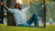 © Yuliia - Middle-aged adult African American businessman in wireless headphones sitting on grass in city park ethnic man listen music in earphones cheerful dancing with hands to song audio funny dance outdoors