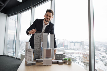 Wall Mural - Confident businessman real estate agents in black stylish formal suit standing while touching 3d model skyscraper of city architecture design at office with panoramic city view.