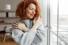 Horizontal Image Of Pretty Redhead Female With Closed Eyes Wearing Rings On Fingers, Hugging Herself, Touching New Soft Sweater, Enjoying Comfort Of Fabric, Standing Next To Panoramic Window At Home