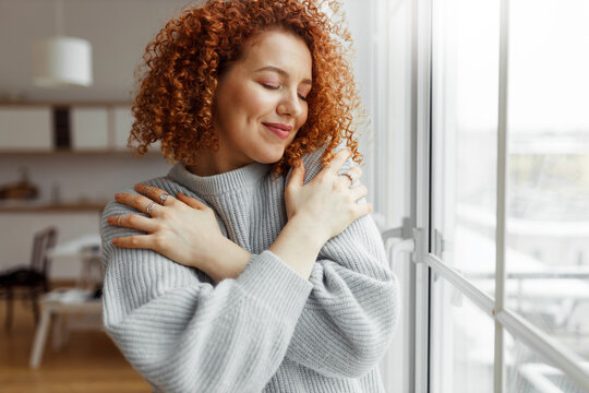 horizontal image of pretty redhead female with closed eyes wearing rings on fingers, hugging herself