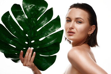 Close up face of beautiful white woman covering her face by green monstera leaf while looking at camera. Female with natural makeup on white isolated background