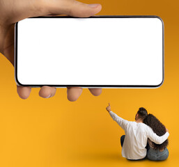 Wall Mural - Couple Hugging Over Yellow Background And Pointing At Huge Smartphone In Hand