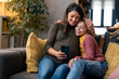 © Dorde - Beautiful smiling mother looking at mobile phone with her lovely daughter while embracing her in living room at home. Mother and daughter using smartphone while resting on couch during the day.