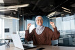 © Liubomir - Portrait of happy smiling and successful muslim at work in office, businesswoman with laptop holding hands up gesture of success and triumph, woman in hijab received online victory notification.