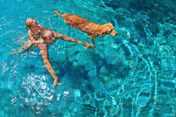 Wall Mural - Mother with little child on back play with fun and train golden labrador retriever puppy in swimming pool. Popular dog like companion, outdoor activity and funny game with family pet on summer holiday