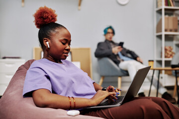 Wall Mural - Side view portrait of young black woman using laptop in office lounge with wireless earphones
