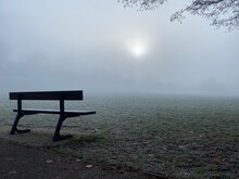 Single Bench In A Park On A Cold, Frosty Morning With The Sun Hidden In The Fog