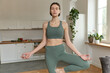 © Folenial - Portrait of a focused relaxed calm young fit brunette woman holding a yoga tree pose while standing in the living room during morning yoga session in a lit living room indoors.