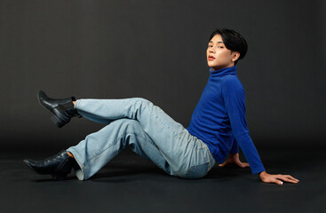 Wall Mural - Portrait studio closeup full body shot of Asian young sexy luxury glamour slim fashionable LGBTQ gay male model in turtleneck long sleeve shirt jeans sitting holding leg up posing on black background