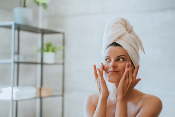 Young adult hispanic woman at bathroom with towel wrapped around head applying cream on face satisfied by spa procedure. Relaxed American female trying new skin care lotion at hotel. Healthy life.