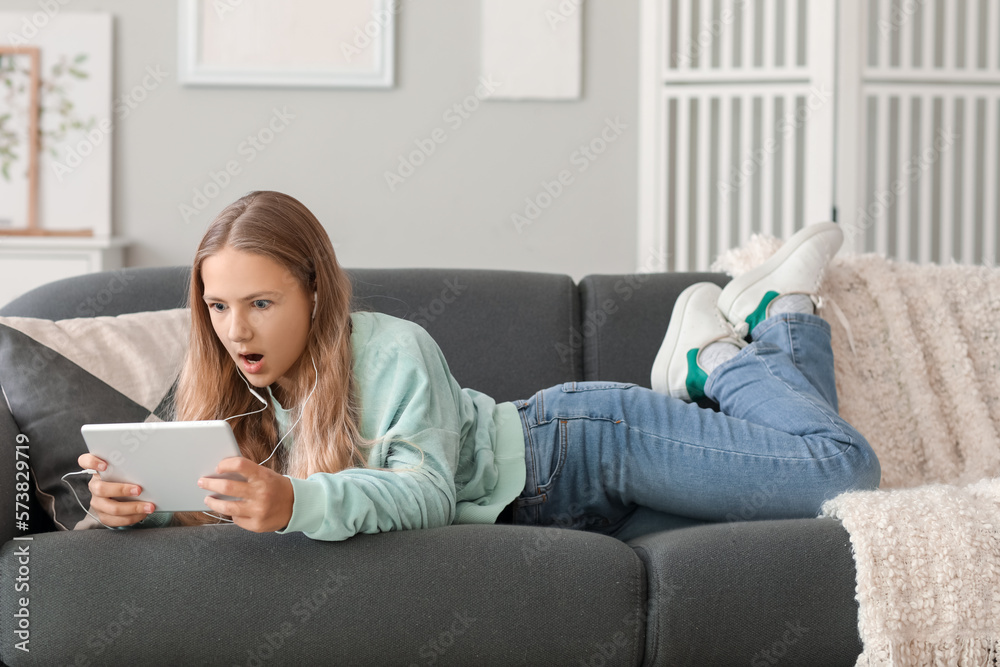 Shocked little girl in earphones using tablet computer on sofa at home