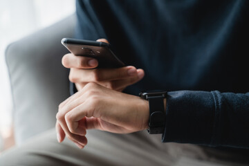 Man using smartphone and smartwatch for tracking activity on sofa in living room at home, wireless connection between the watch and mobile phone.