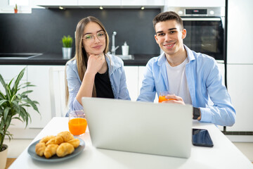 Wall Mural - Portrait of teenage girl and boy in smart casual clothes sitting at desk with laptop and drinking juice at home.