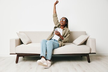 African American woman business freelancer working sitting on the couch at home in the phone, business calls and messages happiness smile, home clothes and eyeglasses, light interior background.