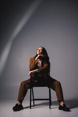 A pretty young woman with a smile on her face and wearing a brown classic suit is sitting on a chair on a gray background. Beautiful woman in a jacket and with makeup on her face