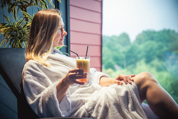 Wall Mural - Woman in white bathrobe lying on sofa and relaxing with orange cocktail at home.