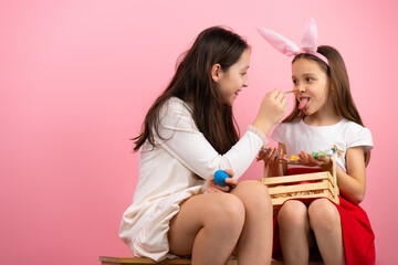 Two lovely sister making fun while preparing for Easter holiday, sitting on a chair with basket and bunny ears and painting the nose.