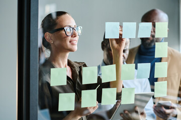 Aufkleber - Young female manager making notes on notepaper while standing in front of transparent board and organizing work against coworkers