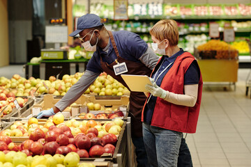 Wall Mural - Mature manager with tablet standing by African American male colleague bending over display with fresh apples while both wearing masks