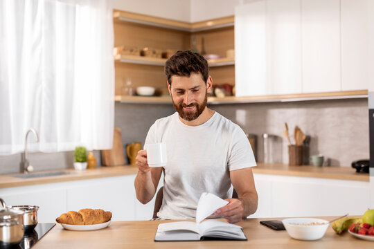Smiling adult caucasian man in white t-shirt enjoy cup of coffee, free time, reading book at table