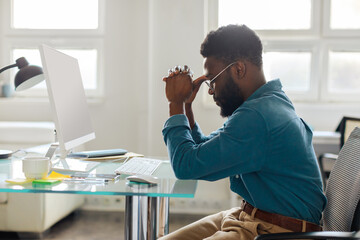 Thoughtful black male entrepreneur in glasses sitting at desk in front of computer, working online in office, side view