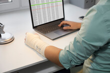 Man With A Hand Injury Working On A Laptop Computer In The Office. Man With A Bandage Wrapped Around His Injured Wrist Sitting His Desk And Using Business Sheets On A Modern Notebook PC
