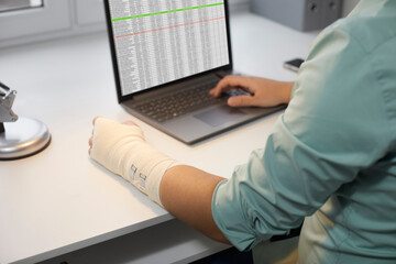 Man with a hand injury working on a laptop computer in the office. Man with a bandage wrapped around his injured wrist sitting his desk and using business sheets on a modern notebook PC