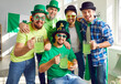 Cheerful friends celebrating St Patrick's Day, having party, drinking beer, and having fun. Group portrait of happy men in green T shirts, hats and glasses holding mugs, looking at camera, and smiling