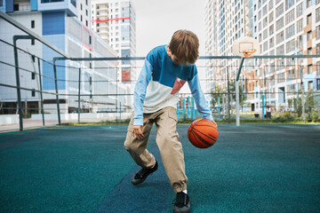 Wall Mural - Active secondary school learner in pants and pullover playing basketball on sports ground after classes or at leisure against houses