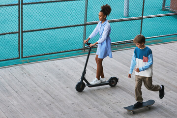 Wall Mural - Two active adolescent children in casualwear riding skateboard and electric scooter while moving along playground surrounded by wire mesh fence